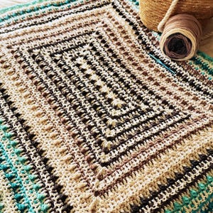 May include: A square, handmade crochet blanket in cream, brown, and teal. A ball of yarn and a crochet hook are next to the blanket. A woven basket with a rope handle is also in the image.