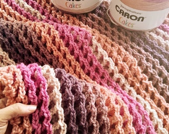 May include: A crocheted blanket with a multi-colored ombre pattern in shades of pink, peach, brown, and beige. The blanket is made with Caron Cakes yarn and has a loose, open weave.