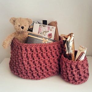 May include: Two crocheted baskets, one larger than the other, filled with various items, including a teddy bear, books, and chocolate. The baskets are a deep burgundy color.