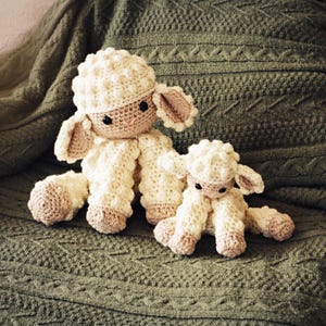 May include: Two handmade crochet sheep toys. The sheep are cream-colored with textured wool-like bodies, tan hooves, and small, tan ears. They have black eyes and are sitting on a green knitted blanket.