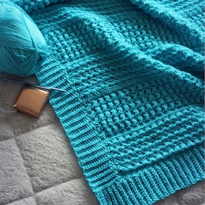 May include: A turquoise blue crocheted blanket with a textured pattern. The blanket is folded and lying on a white surface.