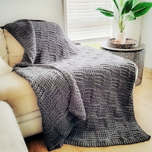 May include: A gray knitted throw blanket draped over a white couch. The blanket has a textured, woven pattern and is made of soft, cozy material.