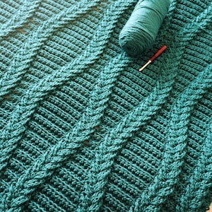 Può includere: Una coperta all'uncinetto color turchese, con un motivo diagonale strutturato. Una palla di filo turchese e un uncinetto rosso sono appoggiati sulla coperta. Il design della coperta presenta sezioni rialzate, intrecciate.