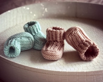May include: Three pairs of knitted baby booties in a light blue and a light brown color. The booties are sitting on a white tray with a textured surface.