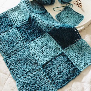 May include: A crocheted blanket made of blue yarn in various shades of blue. The blanket is made of squares and is folded in half. There is a smaller square of the same crochet work on a tray next to the blanket.