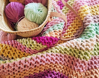 Può includere: Una coperta all'uncinetto colorata con strisce di filato rosa, verde, giallo e arancione. La coperta è realizzata con un punto testurizzato ed è piegata a metà. Un cesto di vimini pieno di gomitoli di lana di vari colori si trova sullo sfondo.