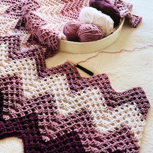 Puede incluir: Una manta de ganchillo hecha a mano en tonos morados y crema, con un patrón en zigzag. Una bandeja ovalada blanca contiene varias madejas de hilo en colores coordinados. También se ven un ganchillo y hilo suelto.