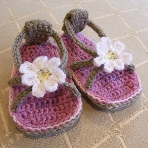 May include: A pair of crocheted sandals with white daisy embellishments. The sandals are pink and brown with a white trim.