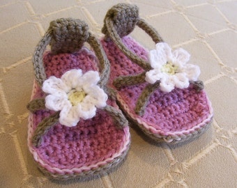May include: A pair of crocheted sandals with white daisy embellishments. The sandals are pink and brown with a white trim.