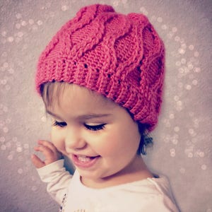 May include: A pink knitted hat with a cable knit pattern. The hat is worn by a child with brown hair and brown eyes.