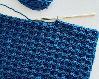 May include: A close-up of a blue crocheted scarf being worked on with a crochet hook. The scarf is made with a textured stitch pattern.
