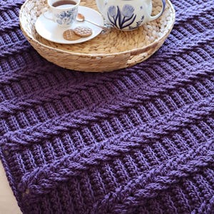 Pode incluir: Um cobertor roxo de crochê com um design texturizado e canelado. Uma bandeja tecida contém uma xícara de chá, um pires, uma colher e biscoitos, juntamente com uma chaleira com detalhes florais azuis. O cobertor está dobrado, mostrando seu padrão intrincado.
