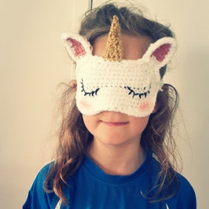 Könnte beinhalten: Eine weiße gehäkelte Schlafmaske in Form eines Einhorns mit rosa Ohren, einem goldenen Horn und gestickten Wimpern.