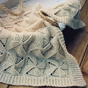 May include: A crocheted baby blanket in a cream color with a delicate, lacy pattern. The blanket is folded and draped over a wicker basket.