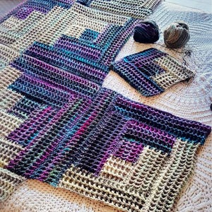 May include: A crocheted blanket in shades of purple, blue, and green, with a textured waffle stitch pattern. The blanket is laid out on a white bedspread, with two balls of yarn and a crochet hook nearby.