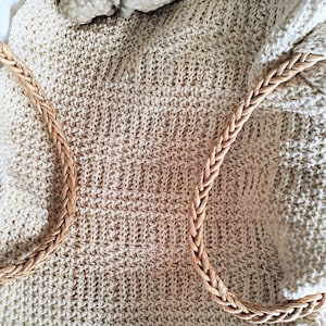 Può includere: Una coperta di maglia color crema con un bordo di corda intrecciata. Due piccole statuine di pecore in ceramica sono posizionate sulla coperta.