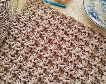 May include: A close-up of a crochet project in progress. The piece is a light brown color with a textured stitch pattern. A ball of yarn, a crochet hook, a measuring tape, and a small gold-colored box are also visible.