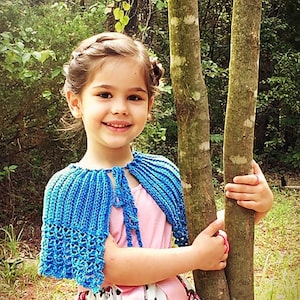 May include: A young girl wearing a blue crocheted cape with a pink shirt and a black and white polka dot skirt. She is standing in front of a tree.