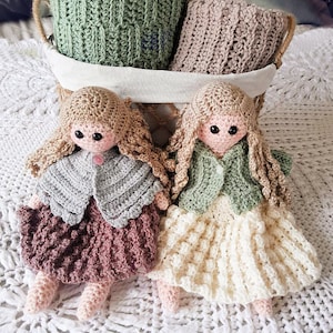 May include: Two crocheted dolls with brown hair, wearing dresses and sweaters, sitting on a white crocheted blanket. The dolls are in a basket with two crocheted blankets.