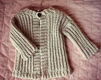 May include: A beige knitted cardigan with a button closure. The cardigan is hanging on a hanger and has a ribbed texture. The text "Child's Play Cardigan" and "design by Lisa van Klaveren" is printed below the cardigan.