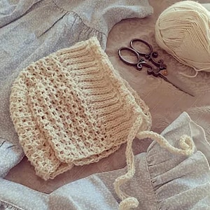 May include: A crocheted baby bonnet in a cream color with a tie string. The bonnet is lying on a light blue fabric with a floral pattern. There is a ball of yarn and a pair of scissors in the background.