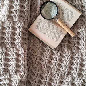 Puede incluir: Una manta de crochet beige con un libro y una lupa encima. El libro está abierto y la lupa está apoyada en las páginas.