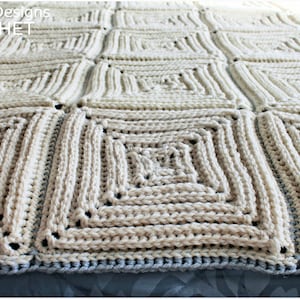 May include: A close-up of a crocheted blanket with a square pattern in a cream color. The blanket has a blue border.