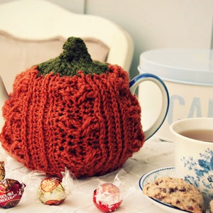 Puede incluir: Un calienta-tetera de punto de ganchillo en forma de calabaza de color naranja y verde, sobre un mantel blanco con una taza de té, un platillo, galletas y chocolates.