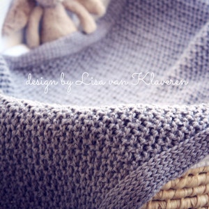May include: A gray knitted blanket with a textured pattern is draped over a woven basket. A stuffed bunny is nestled within the blanket. The text "design by Lisa van Klaveren" is visible on the blanket.