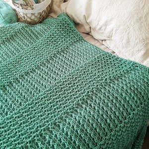 May include: A teal crocheted throw blanket on a white bed with a basket of yarn in the background.