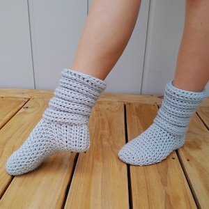 Puede incluir: Un par de calcetines de pantuflas de crochet gris claro con un puño doblado. Los calcetines están sobre un suelo de madera.
