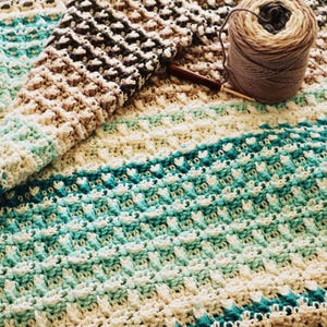 Pode incluir: Um cobertor de crochê feito à mão em tons de branco, verde-azulado, castanho e bege. O cobertor apresenta um padrão de ponto texturizado e está parcialmente dobrado, revelando o design intrincado. Uma bola de lã e uma agulha de crochê também são visíveis.