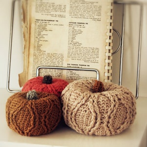 May include: Three crocheted pumpkins in different colors and styles. The pumpkins are sitting on a white surface with a vintage cookbook in the background. The cookbook has recipes for "Frost-on-the-Pumpkin Pie", "Cavalier Pumpkin Pie", and "Old Fashion Pumpkin Pie".