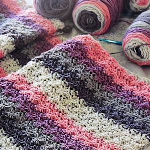 May include: A striped crocheted blanket in shades of pink, purple, grey, and white. The blanket is made with chunky yarn and has a textured stitch pattern.