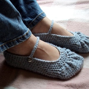 May include: A pair of light blue crocheted slippers with a button detail on the strap. The slippers are worn by a person wearing blue jeans.