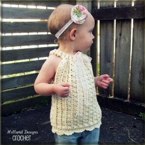 May include: A young child wearing a crocheted halter top with a white lace trim and a floral headband. The top is a light cream color and the headband has a pink and green flower.