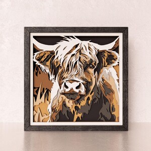 May include: A square wall art piece featuring a layered, multi-dimensional cutout of a Highland cow. The artwork uses shades of brown, cream, black, and white to create depth and texture, all enclosed within a dark, textured wooden frame.