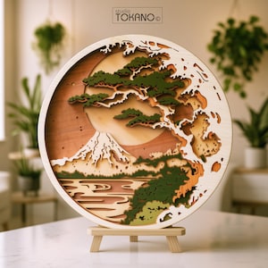 Puede incluir: Una pieza de arte mural circular de madera multicapa que presenta un paisaje estilizado con una montaña, un gran sol, un pino y olas. La obra se exhibe sobre un pequeño soporte de madera e incluye el texto studio TOKANO en la parte superior.