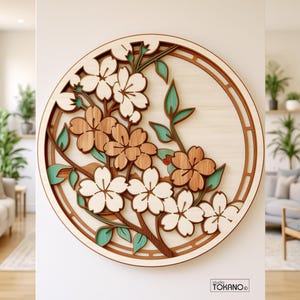 Op de afbeelding: Een cirkelvormig houten wandkunstwerk met gelaagde kersenbloesemtakken, witte en naturelkleurige bloemen en groenblauwe bladeren. Het ontwerp is gevat in een ronde lijst met een uitgesneden patroon. De tekst studio TOKANO is zichtbaar.