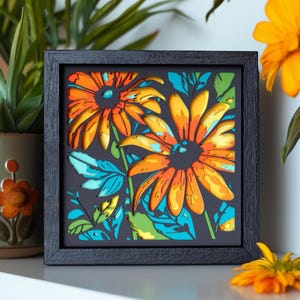 May include: A square, black-framed wall art piece featuring a multi-layered, laser-cut design of two large orange and yellow flowers with teal and green foliage accents. The artwork displays depth through stacked layers against a dark background.