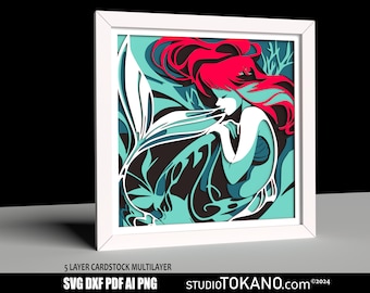May include: A 5-layer cardstock multi-layer papercraft design of a mermaid with flowing red hair and a teal tail. The design is framed in white and features intricate details.