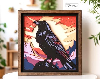 May include: A square wooden framed wall art piece featuring a multi-layered paper cutout of a black raven perched on a rock. The background displays a sunset with shades of red, orange, and yellow, with purple mountains and trees below.