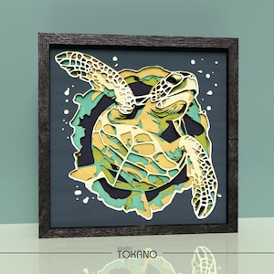 May include: A multi-layered paper art piece featuring a sea turtle in shades of yellow, green, and teal, set against a dark blue background within a square black frame. The text "studio TOKANO" is printed in black at the bottom of the frame.