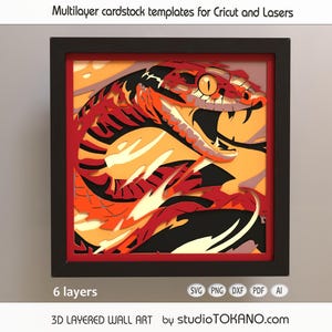 May include: A framed 3D layered wall art piece depicting a snake. The artwork uses multiple layers of cardstock in red, orange, yellow, and black. The frame is black, and the piece is labeled "6 layers". The text "Multilayer cardstock templates for Cricut and Lasers" is at the top.