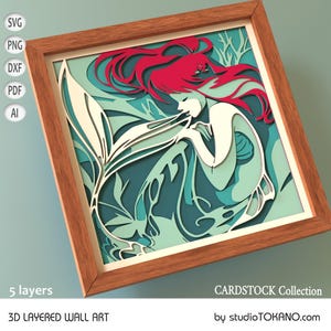 Op de afbeelding: Een 3D gelaagd wandkunstwerk met een zeemeermin met rood haar en een witte staart. De zeemeermin is omringd door turquoise en groen gebladerte. De kunst is ingelijst in een houten lijst en heeft 5 lagen. CARDSTOCK Collection van studioTOKANO.com