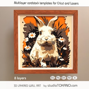May include: A framed 3D layered wall art piece featuring a white rabbit surrounded by daisies and foliage. The artwork is composed of multiple layers, creating a dimensional effect. The frame is brown. The text "Multilayer cardstock templates for Cricut and Lasers" is at the top.