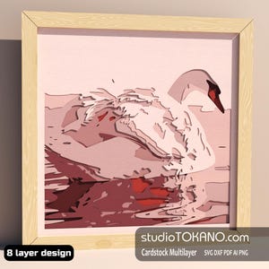 May include: A framed paper art piece featuring a swan in shades of pink and red. The artwork, with a light wood frame, shows a swan on water. Text includes "studioTOKANO.com" and "8 layer design".