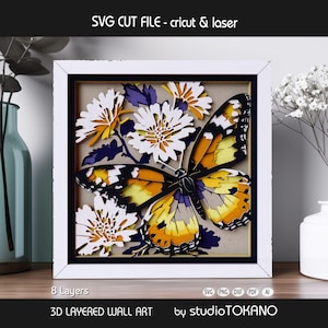 May include: A framed 3D layered wall art piece featuring a butterfly and flowers. The butterfly has orange, yellow, black, and white wings. The flowers are white and yellow. The artwork is in a white frame with the text "SVG CUT FILE - cricut & laser" and "3D LAYERED WALL ART".