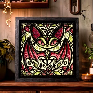 May include: A square framed wall art piece featuring a multi-layered paper cut design of a stylized bat. The artwork uses a color palette of black, cream, red, and light green, with intricate symmetrical patterns and cutouts creating depth.