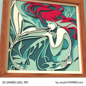 Op de afbeelding: Een 3D gelaagd wandkunstwerk met een zeemeermin met golvend rood haar, een witte staart en een turquoise en groene achtergrond. De kunst is ingelijst in een natuurlijk houten frame.